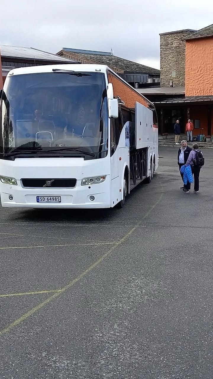 Hvit turbuss fra Frolandsruta parkert foran en bygning, en reisende på vei forbi